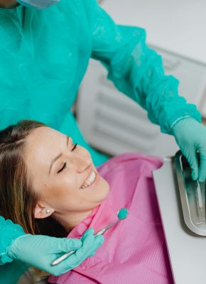 A woman sits smiling in a dental chair while a dentist prepares instruments.