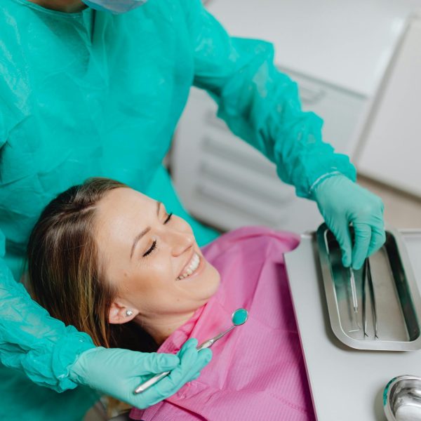 A woman sits smiling in a dental chair while a dentist prepares instruments.